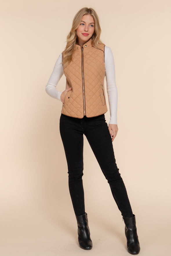 Blonde woman wearing zipped up tan vest over long sleeve white shirt and black jeans with black leather booties