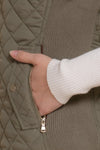 Close up of a hand in an olive green vest pocket and white long sleeve shirt