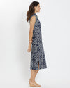 Brunette woman facing sideways with hands behind her back wearing geometric print navy and white rope print midi dress with tan wedge sandals