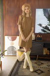 Blonde woman in brown camel colored tweed dress with braided neckline and pockets with gold buttons, wearing matching tan felt hat and light beige boots standing in front of wooden paneled wall with artwork