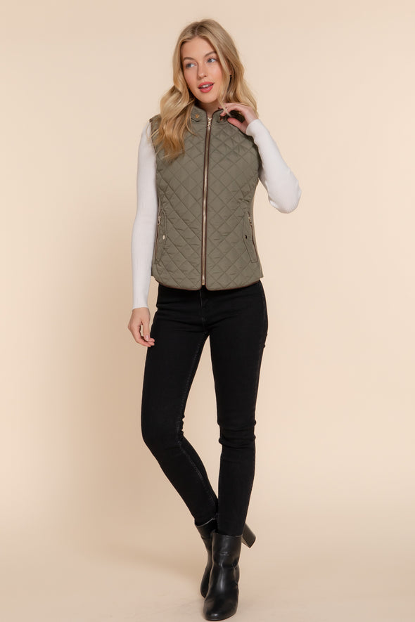 Blonde woman wearing zipped up olive green vest over long sleeve white shirt, black skinny jeans and black ankle boots